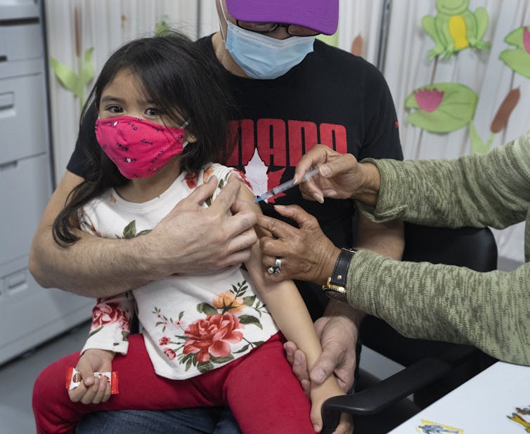 A girl in a pink face mask sits in a man's lap as she's vaccinated.THE CANADIAN PRESS/Ryan Remiorz
