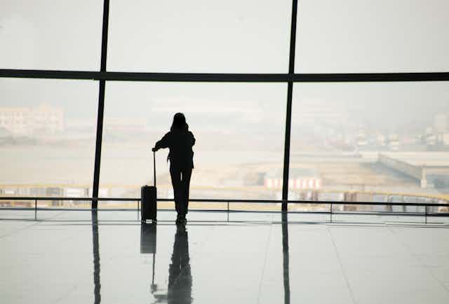 silhouette of a woman leaning on the handle of her luggage looking out the window of an airport terminal.