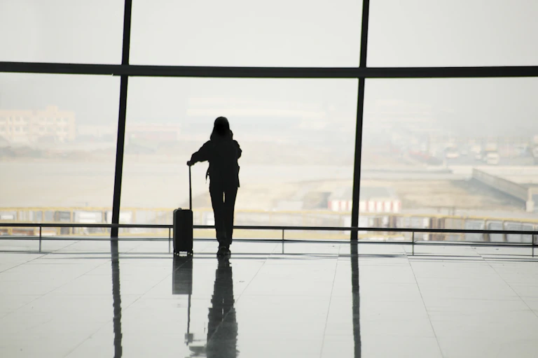 silhouette of a woman leaning on the handle of her luggage looking out the window of an airport terminal.