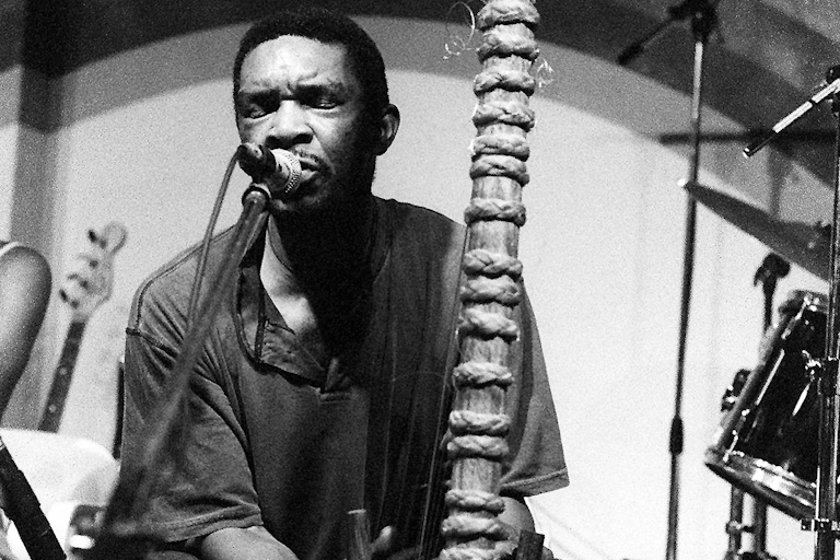 Black and white image of a man playing the strings of a wooden musical instrument, singing into a microphone alongside it, his eyes closed.