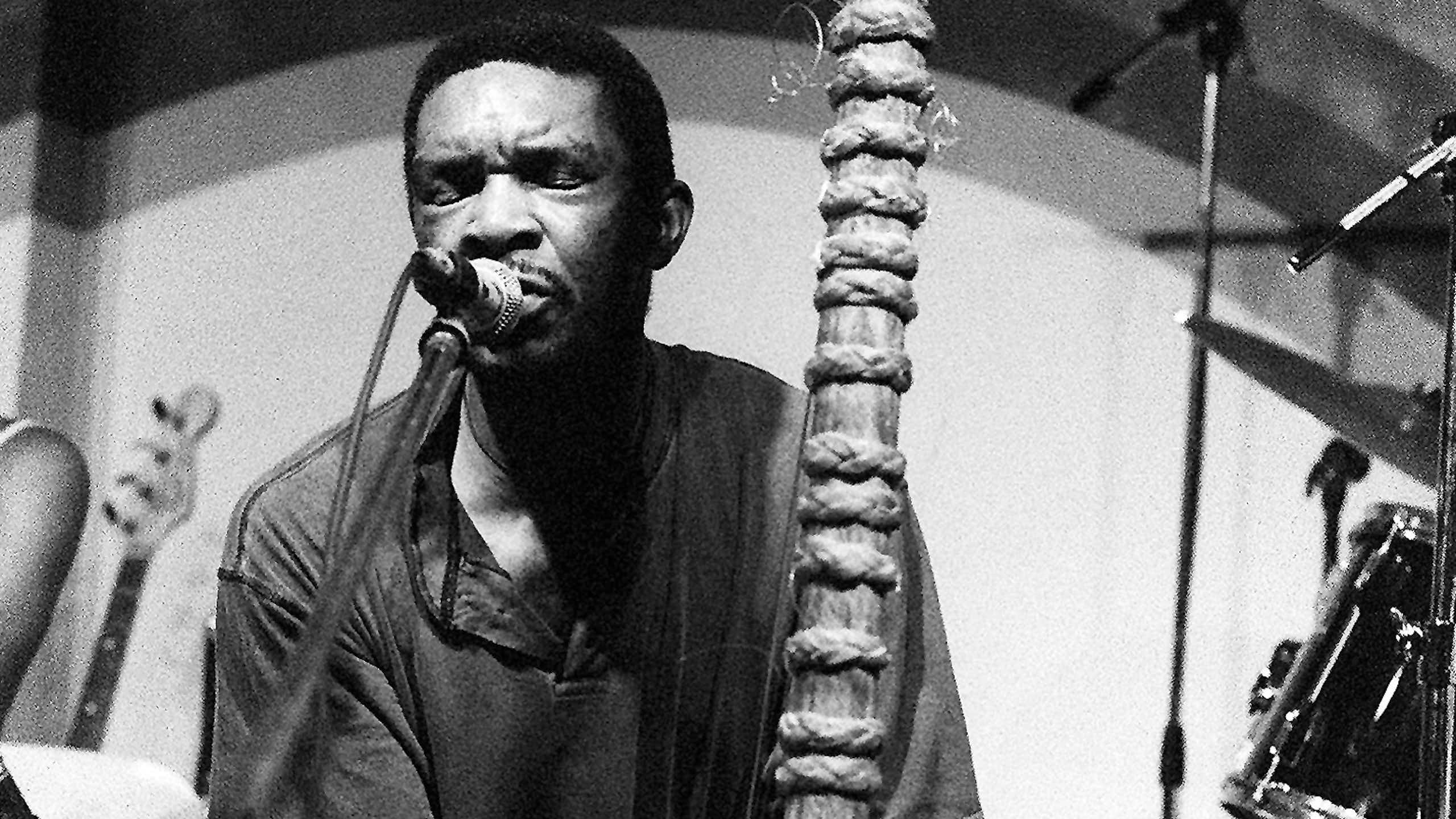 Black and white image of a man playing the strings of a wooden musical instrument, singing into a microphone alongside it, his eyes closed.