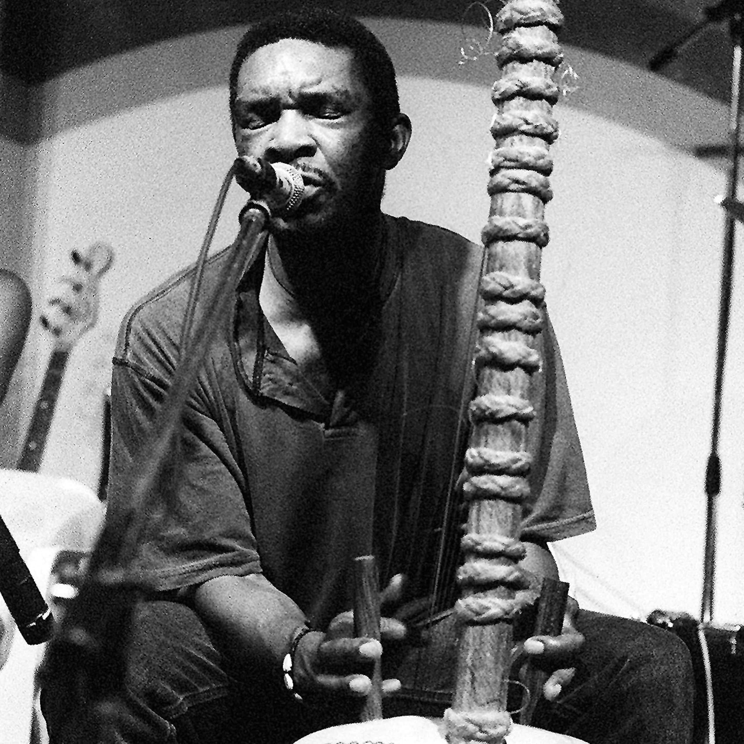 Black and white image of a man playing the strings of a wooden musical instrument, singing into a microphone alongside it, his eyes closed.