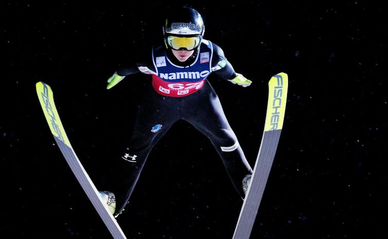 A ski jumper in the air after a jump wearing googles and helmet.
