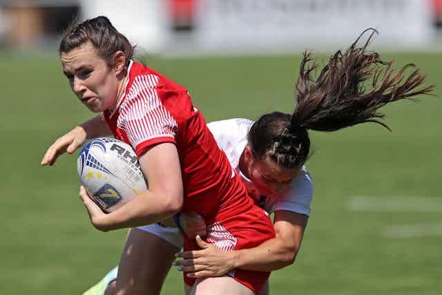 A woman rugby player wearing red, being tackled.