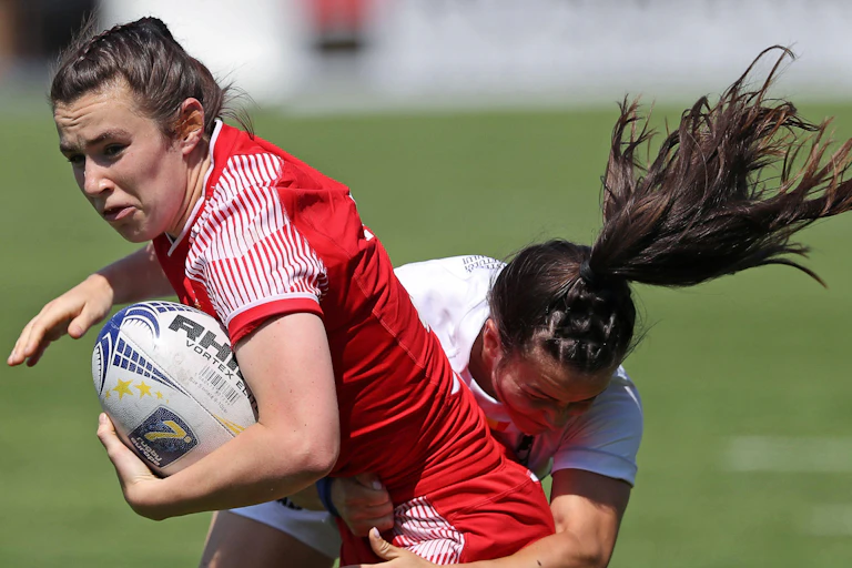 A woman rugby player wearing red, being tackled.