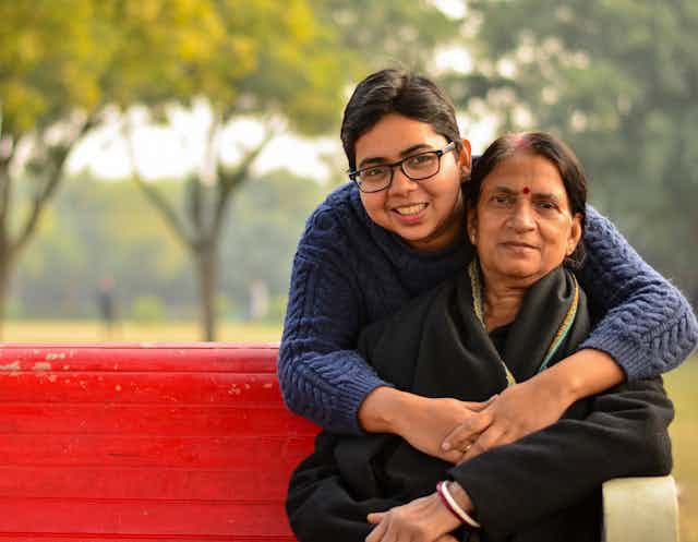 A young South Asian woman hugging an older South Asian woman on a red park bench
