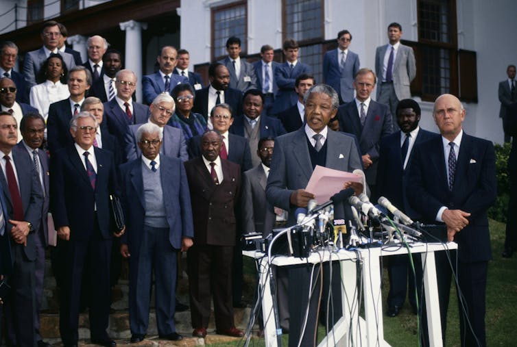 A Black man and a white man stand in front of a group of other men while answering questions from reporters.