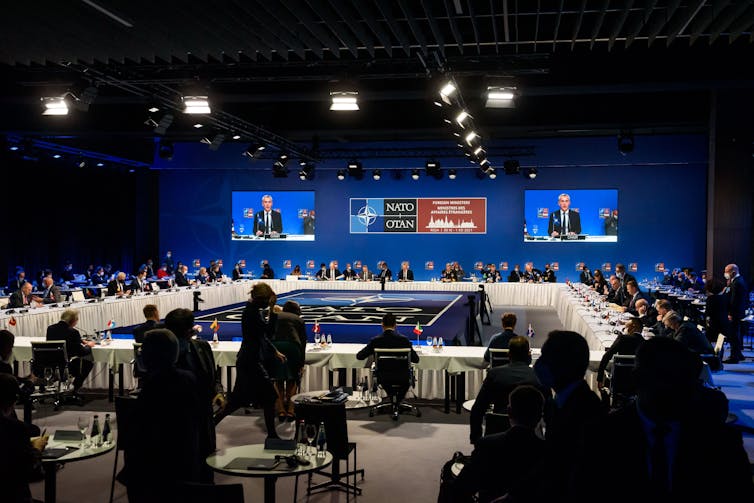 A room full of people sit around a table watching several TV screens.