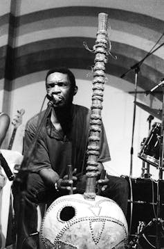 A man plays a traditional music instrument with strings and a long neck, leaning into a microphone on a stage.