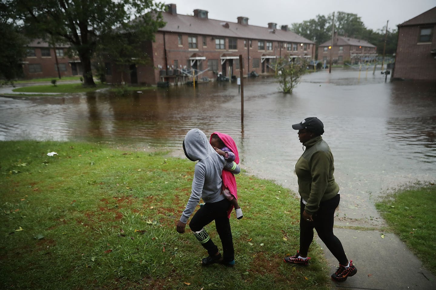 A woman carries a child past an area where flood water surrounds low-rise apartment buildings.