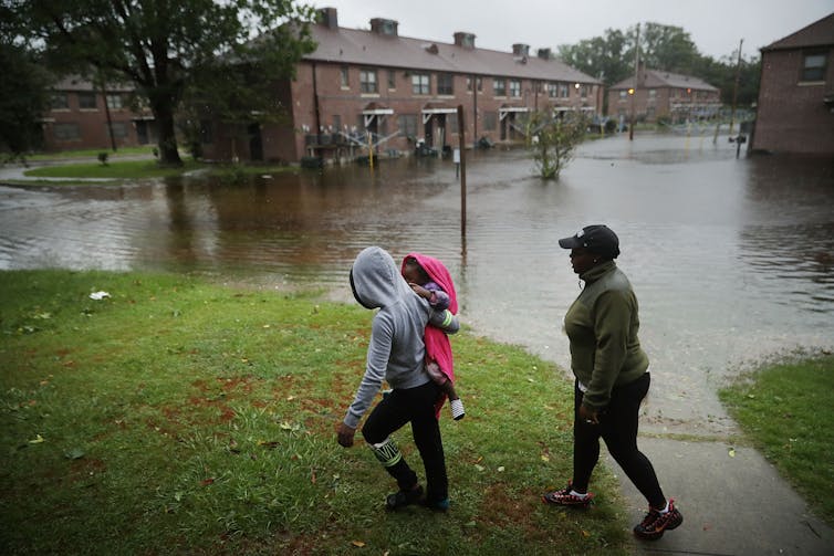 A woman carries a child past an area where flood water surrounds low-rise apartment buildings.