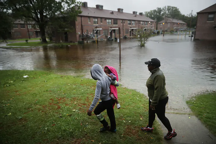 A woman carries a child past an area where flood water surrounds low-rise apartment buildings.