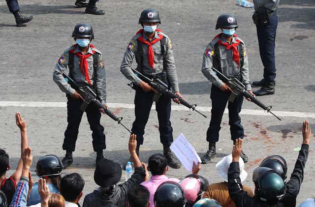 Three armed anti-riot police wearing red neckechiefs stand side by side, facing protesters in the foreground. Many are lifting three finger symbols of resistance.