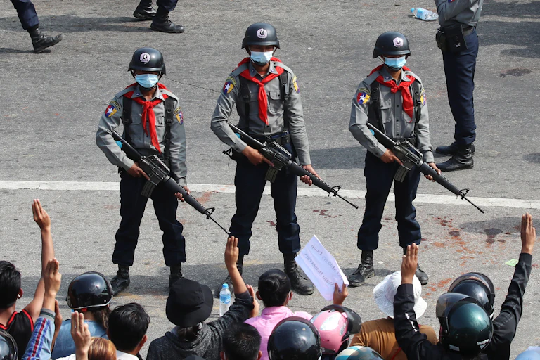 Three armed anti-riot police wearing red neckechiefs stand side by side, facing protesters in the foreground. Many are lifting three finger symbols of resistance.