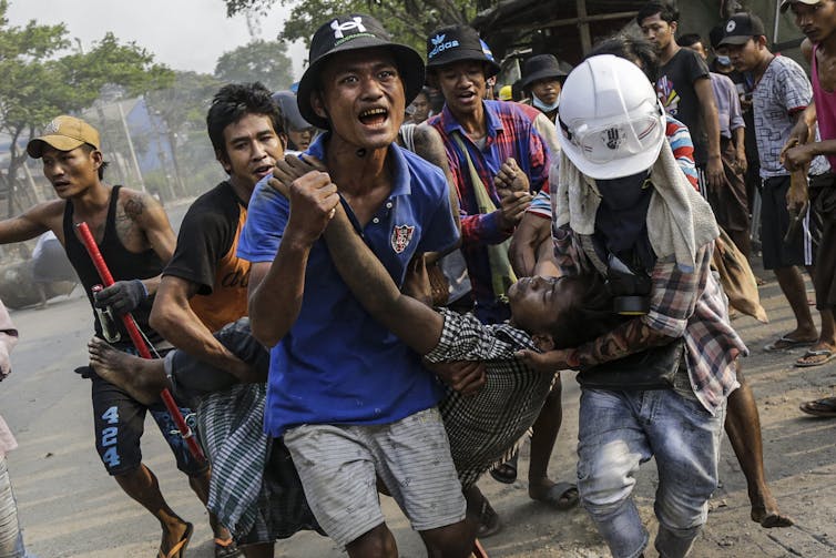 Group of Burmese men carry injured man while shouting.