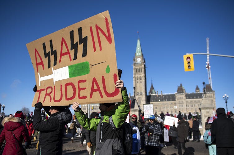 A sign reads Assassin Trudeau amid a group of protesters with Parliament buildings in the background.
