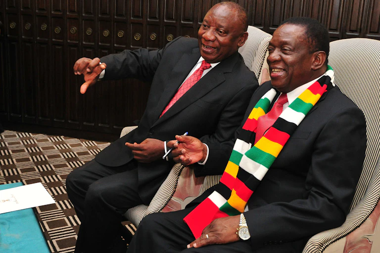 Two men wearing suits smile as one illustrates a point to the other with his right hand while they're both seated.