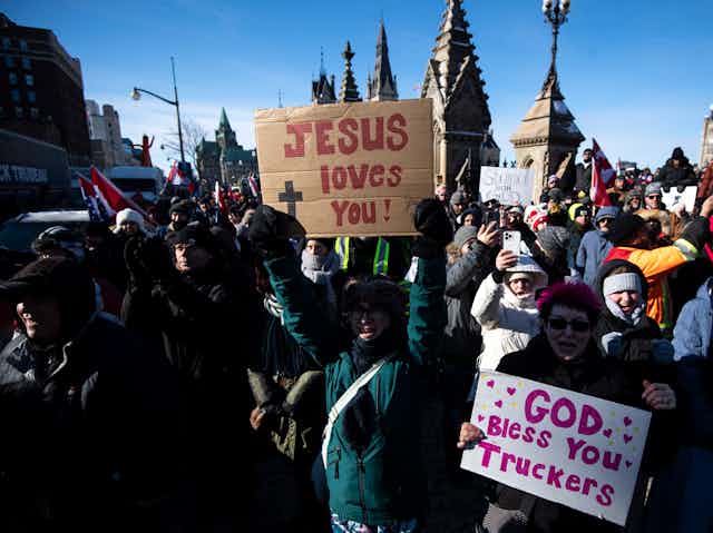 People hold signs reading Jesus Loves You and God Bless You Truckers with Parliament buildings behind them.