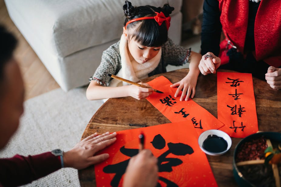 A Chinese girl, sitting at a table, with grandmother by her side,learns how to write Chinese New Year auspicious messages in Chinese calligraphy.