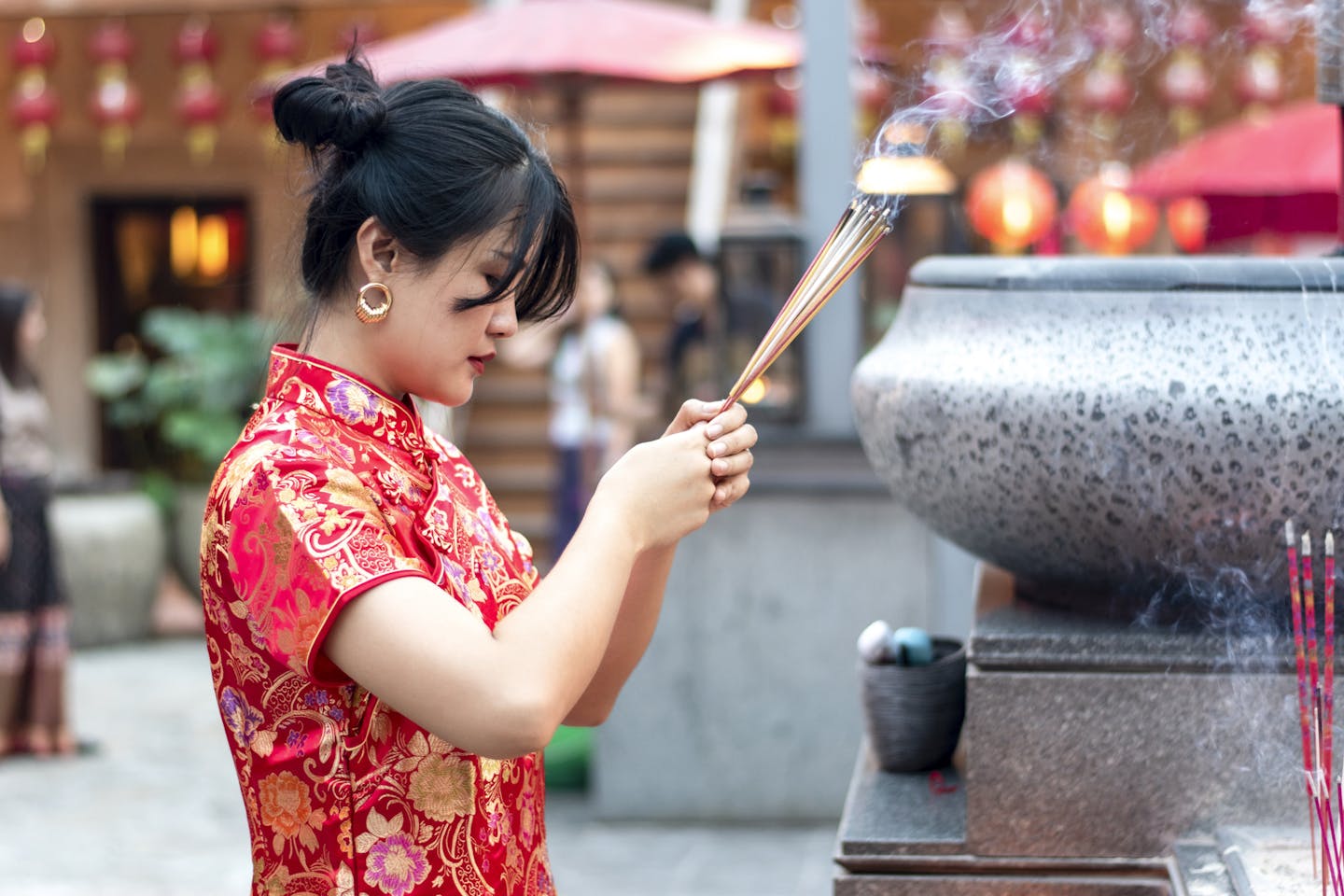 A woman dressed in a festive red Chinese dress holds incense sticks, with a bowed head, as she offers them to a deity.