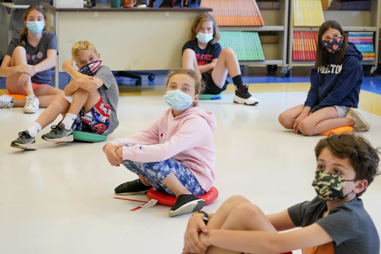 Children wearing masks sit on a classroom floor