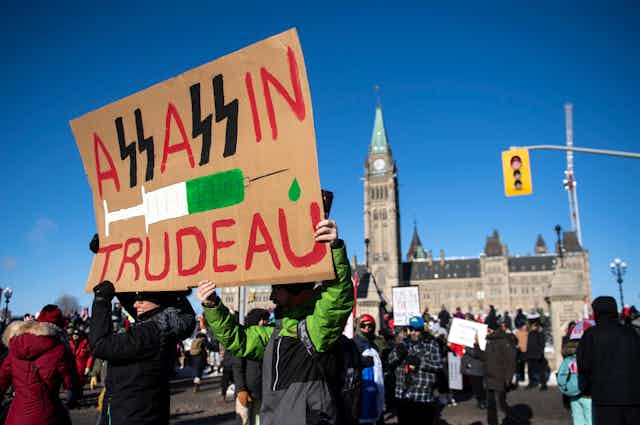 In front of Parliament Hill, two people hold a sign reading 'Assassin Trudeau' but the letters S are replaced with Nazi SS.