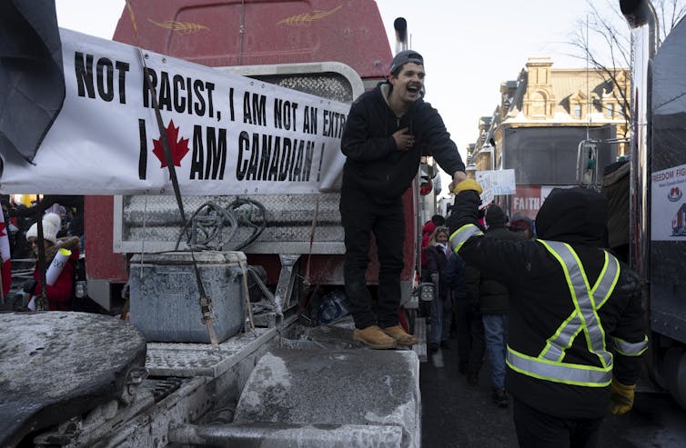 A man stands in front of a sign on a transport truck that reads 'I am not a racist, I am not an extremist, I am Canadian'