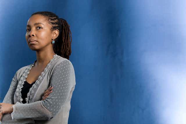 A woman stands against a blue backdrop, her arms folded, a serious expression on her face.