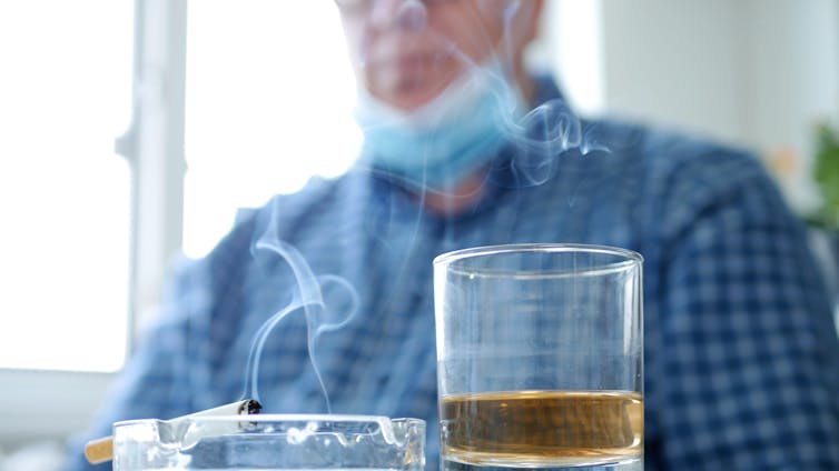 a short glass drink with alcohol next to an ashtray with a smoldering cigarette in it. in the background, out of focus, a man wearing a blue shirt and surgical mask