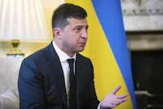 A man in a dark suit gestures and talks with a blue and yellow flag and a table lamp behind him.
