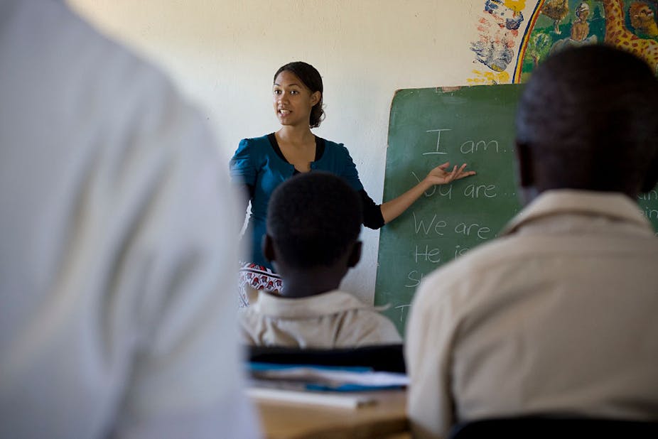 A young teacher pointing towards the chalkboard and facing her pupils in a classroom.