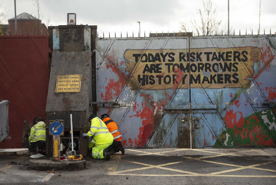 Three workers in high-vis kneeling beside an imposing gate painted with the phrase Today's Risk Takers are Tomorrow's History Makers.