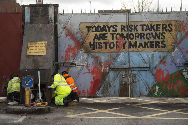 Three workers in high-vis kneeling beside an imposing gate painted with the phrase Today's Risk Takers are Tomorrow's History Makers.