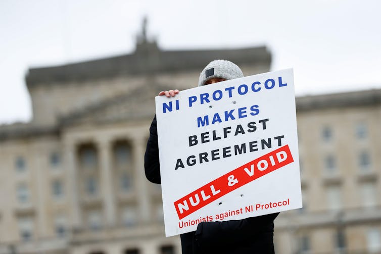 A man holds a sign reading NI Protocol Makes Belfast Agreement Null and Void. The parliament building in Belfast can be seen behind him.