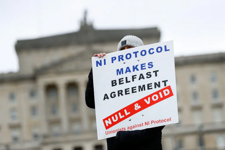 A man holds a sign reading NI Protocol Makes Belfast Agreement Null and Void. The parliament building in Belfast can be seen behind him.
