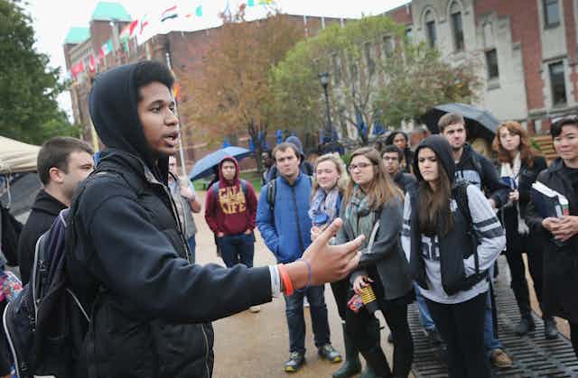 A Black man gesticulates to a crowd of mostly white students on a college campus