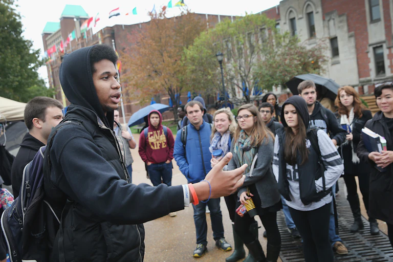 A Black man gesticulates to a crowd of mostly white students on a college campus