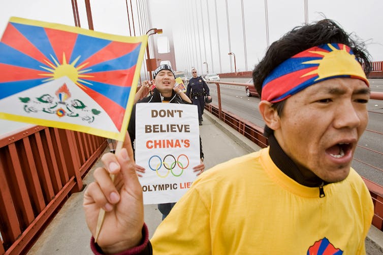 A man in a yellow shirt holds up a Tibetan flag, marching in front of a person who has a sign that says 'Don't believe China's Olympic lies.' They walk across the Golden Gate Bridge in San Francisco