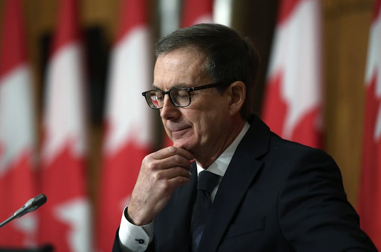 A man wearing glasses at a press conference with a row of Canadian flags behind him.