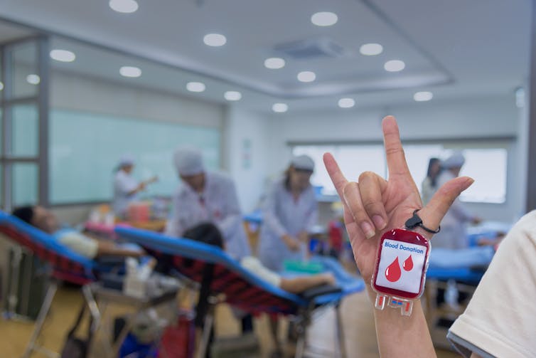 A person holds an I love you sign and a small blood bag, with nurses and blood donors in the background.