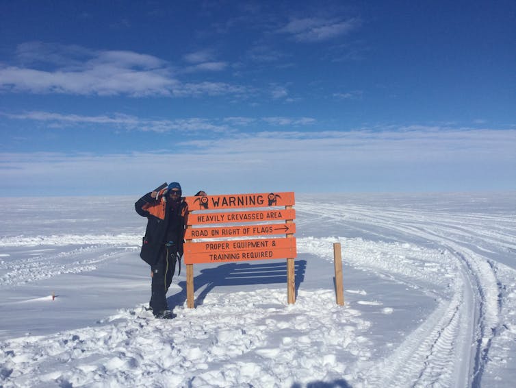 A person next to a sign, and tracks across the ice, during a trip across the Ross Ice Shelf