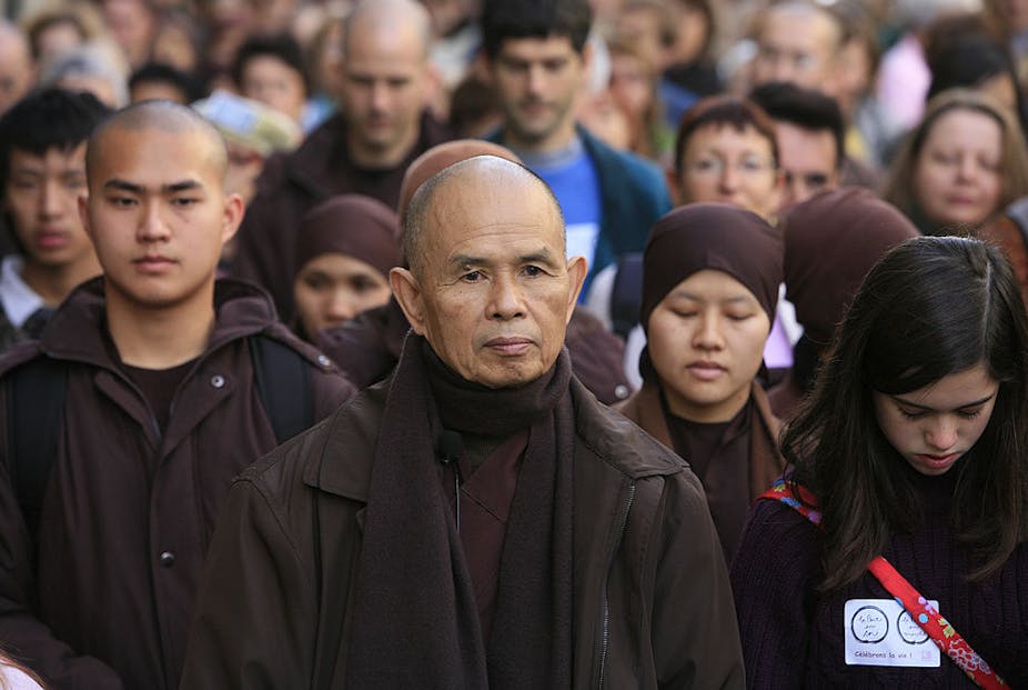 A monk stands, looking pensive, at the front of a crowd of people.
