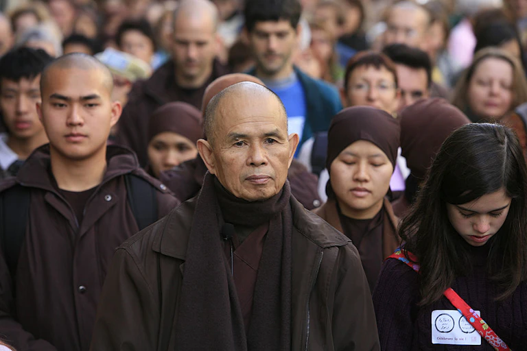 A monk stands, looking pensive, at the front of a crowd of people.