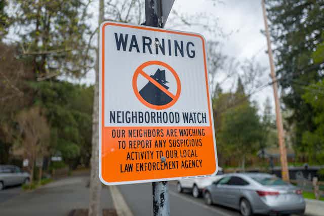 An orange and white sign in a leafy area, cars passing. The sign reads 'Neighbourhood watch' with an illustration of a shady looking criminal figure crossed out.
