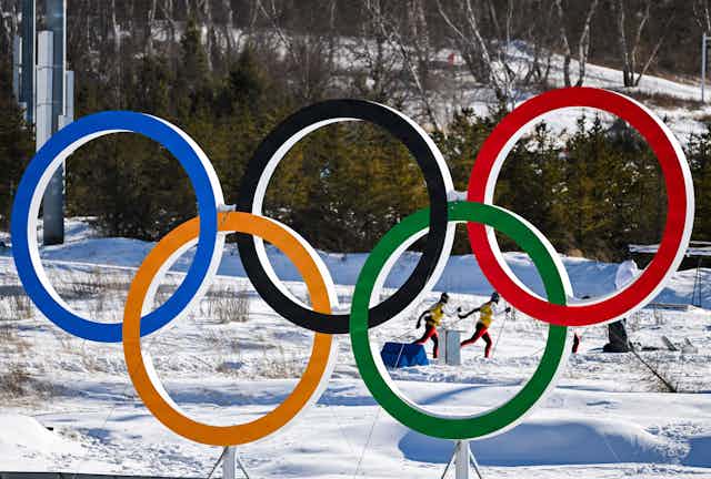 Two cross country skiers practicing on a snowy field behind a large sculpture of the Olympic rings.