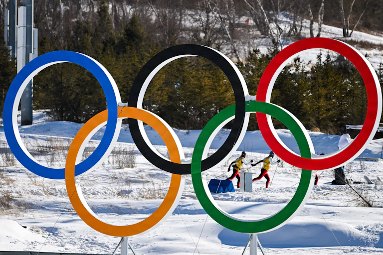 Two cross country skiers practicing on a snowy field behind a large sculpture of the Olympic rings.