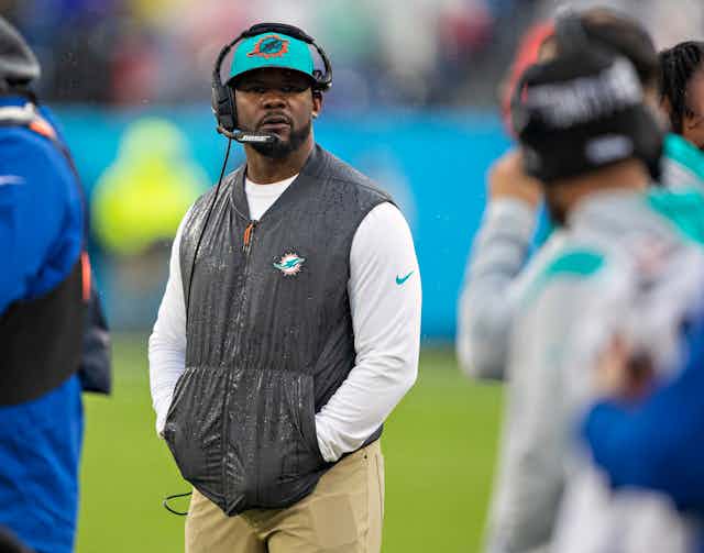 A Black man wearing a headset on the sidelines of a football game.