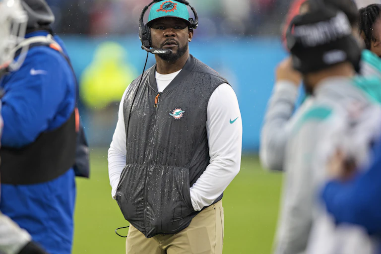 A Black man wearing a headset on the sidelines of a football game.
