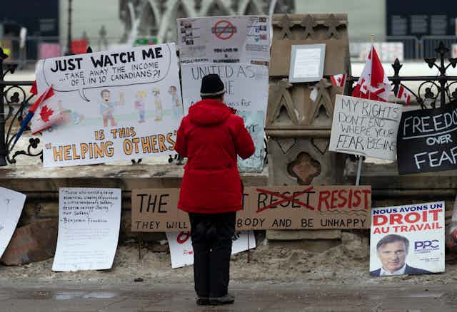 A woman in a red coat takes a photo of a collection of signs attached to a fence.