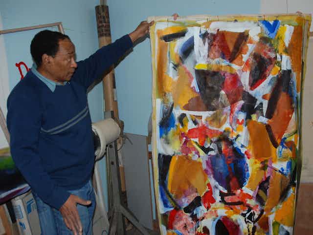 A man dressed in a navy jersey and jeans looks earnestly at a large canvas that he holds with his left arm, draped on a frame. It shows an abstract artwork of reds, oranges and blues, shapes merging and covering the canvas.